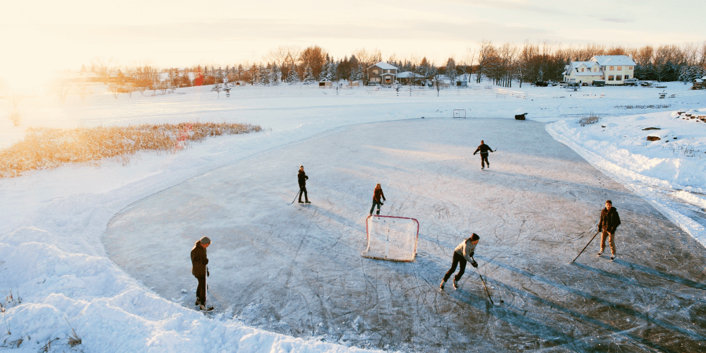 Group of people playing ice hockey on a frozen pond near residential houses during sunset.