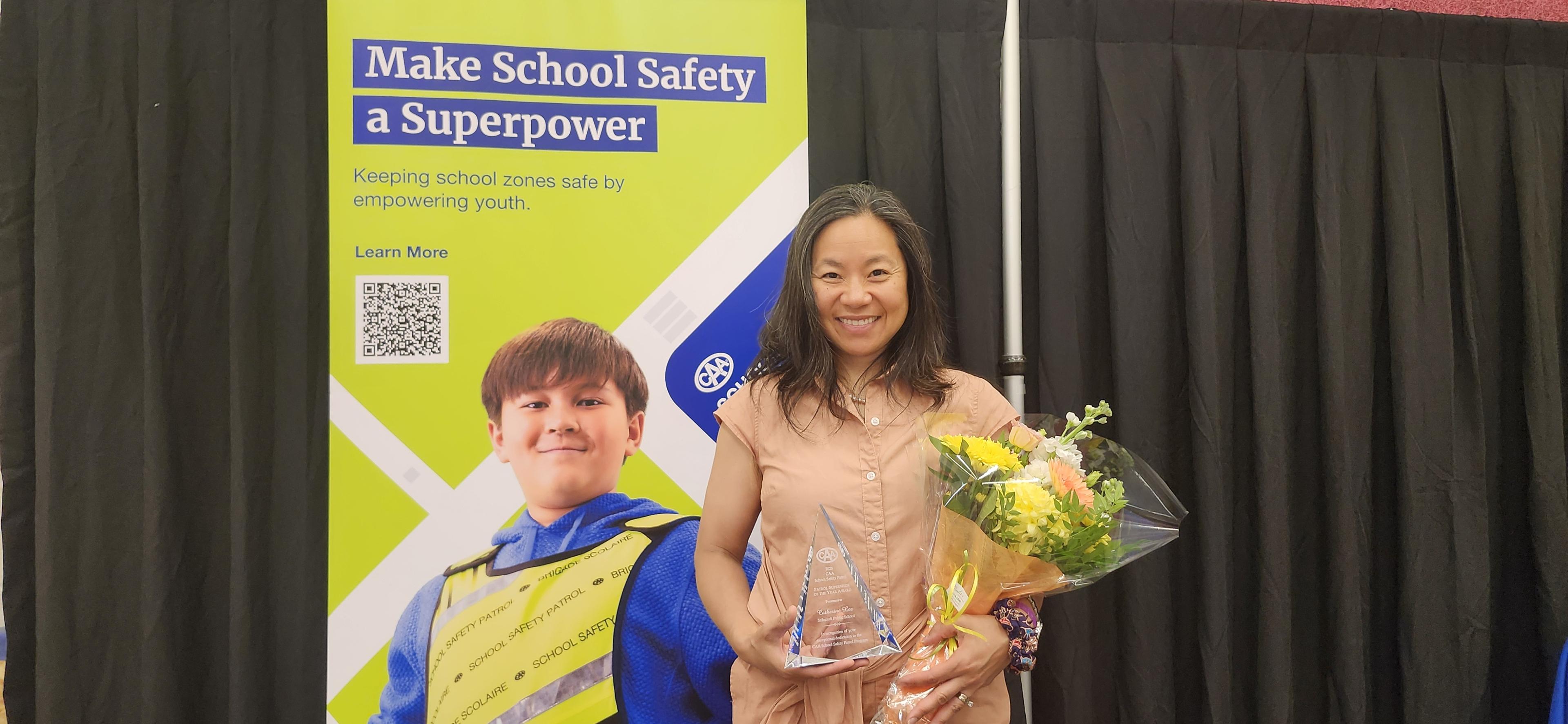 A woman stands in front of a banner holding an award and a bouquet of flowers.