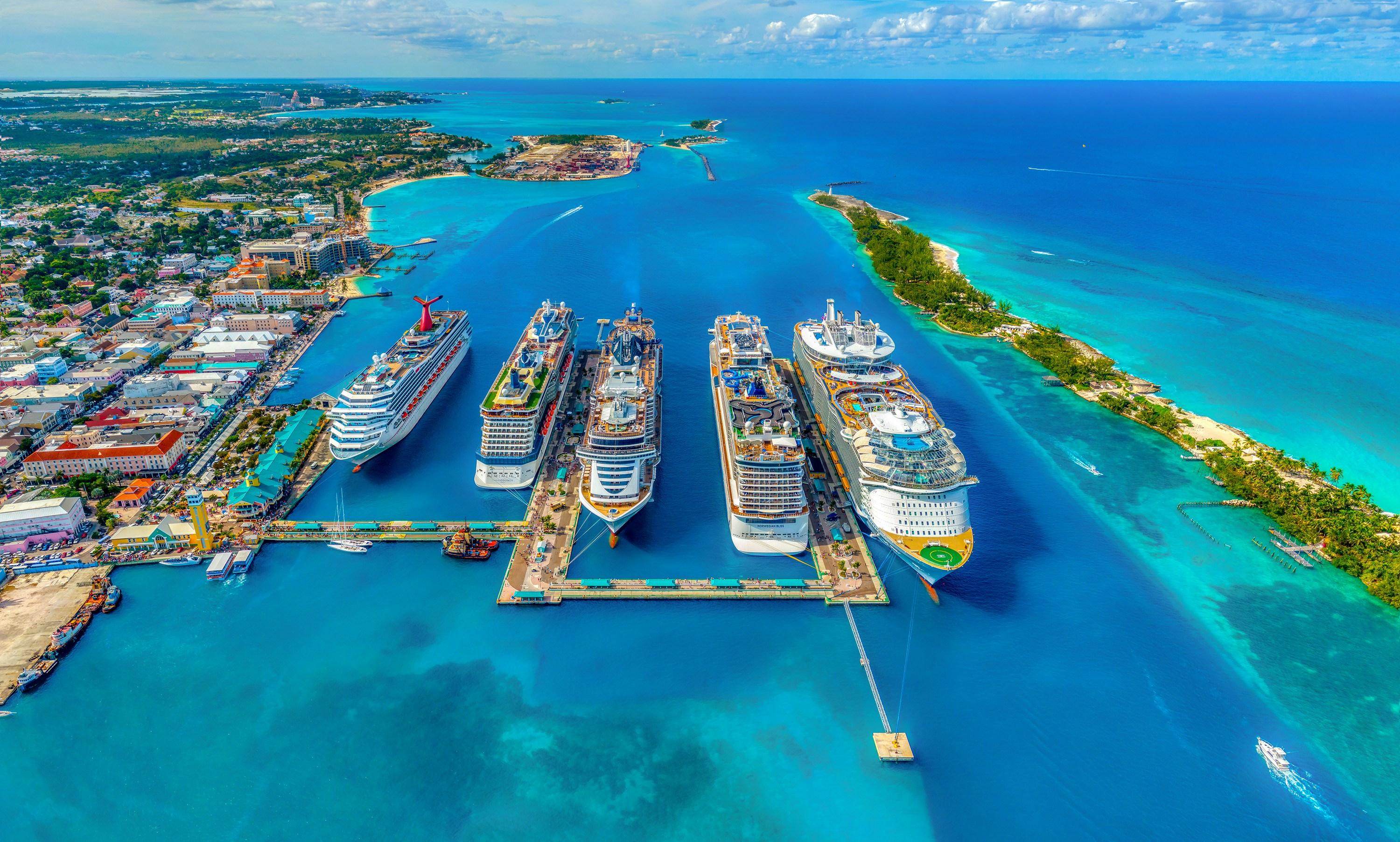 An overhead shot of a cruise ship port. There are five cruise ships docked in the port, floating in bright blue water.