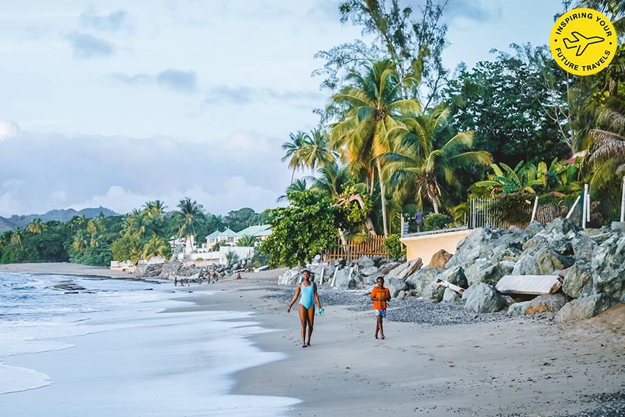 Two people walking on a tropical beach with palms and residential buildings in the background.