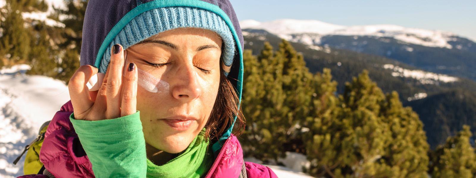 A woman wearing colourful winter gear stands outdoors with snow-capped mountains behind her. Her right arm is raised as she applies a white cream to her face.