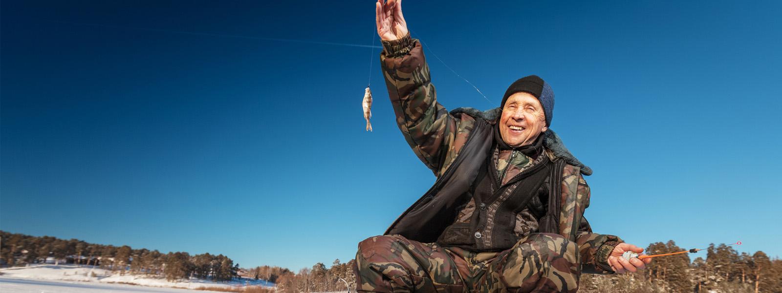 A man wearing camouflage-printed winter gear sits on a stool on a frozen lake. In one hand, he holds a thin fishing rod, and in the other he holds up a fishing line — at the end of it s a small silver fish.