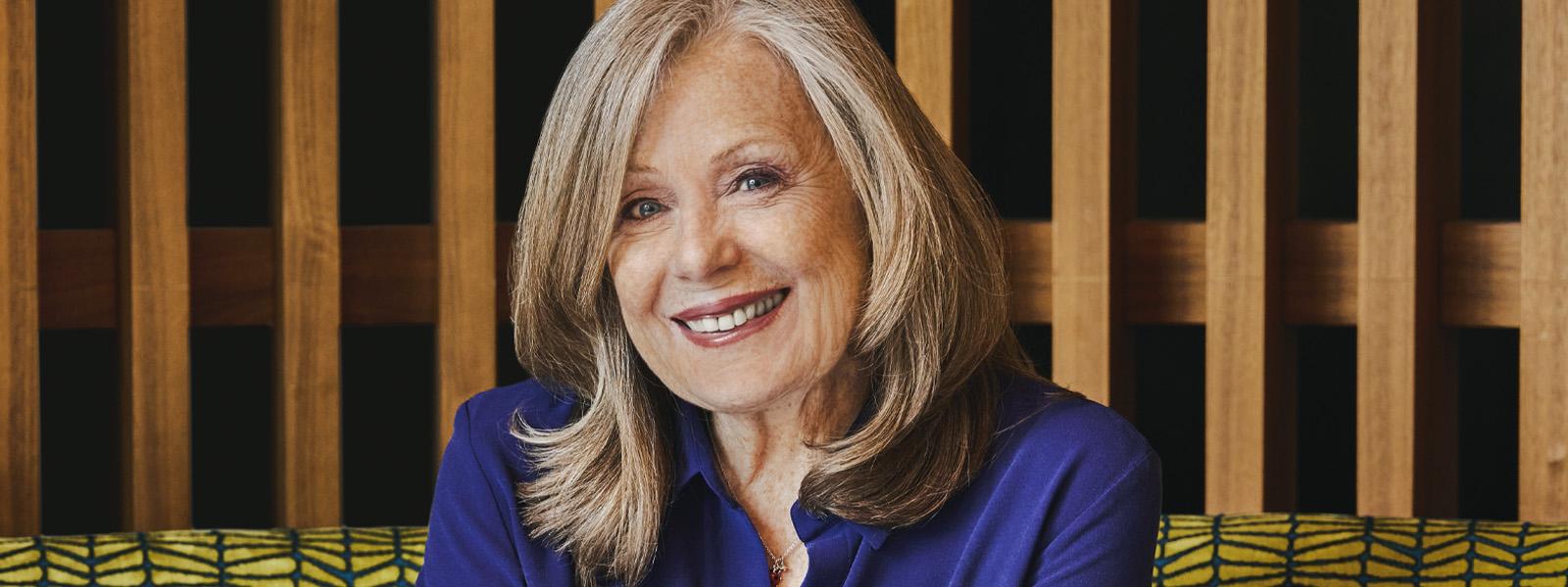 A woman with short, grey hair sits on a green couch with wooden slats on the wall behind her. She wears a blue blouse and smiles at the camera.