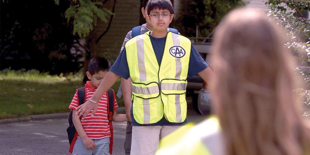 A student wearing a safety vest assists a younger child crossing the street as part of a school safety patrol program.