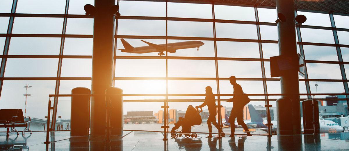 A family, two parents and a child in a stroller, walk through an airport. Behind them is a large window, showing a plane taking off.