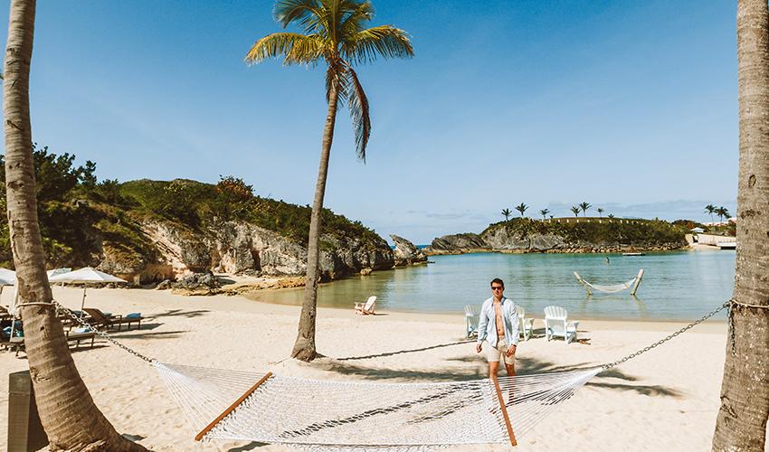 Man walking on a tropical beach with palm trees, hammocks, and sunbathers in the background.