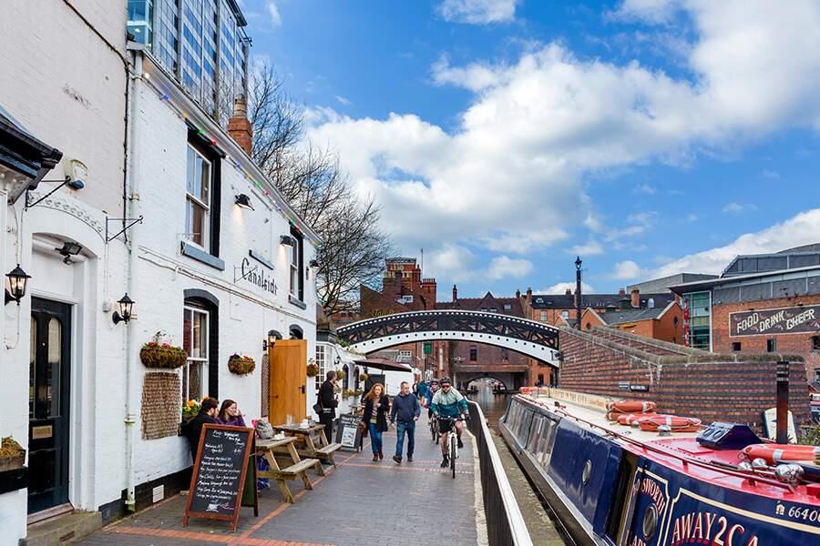 A bustling canal side with people walking and cycling, traditional buildings, and moored boats under a blue sky with clouds.