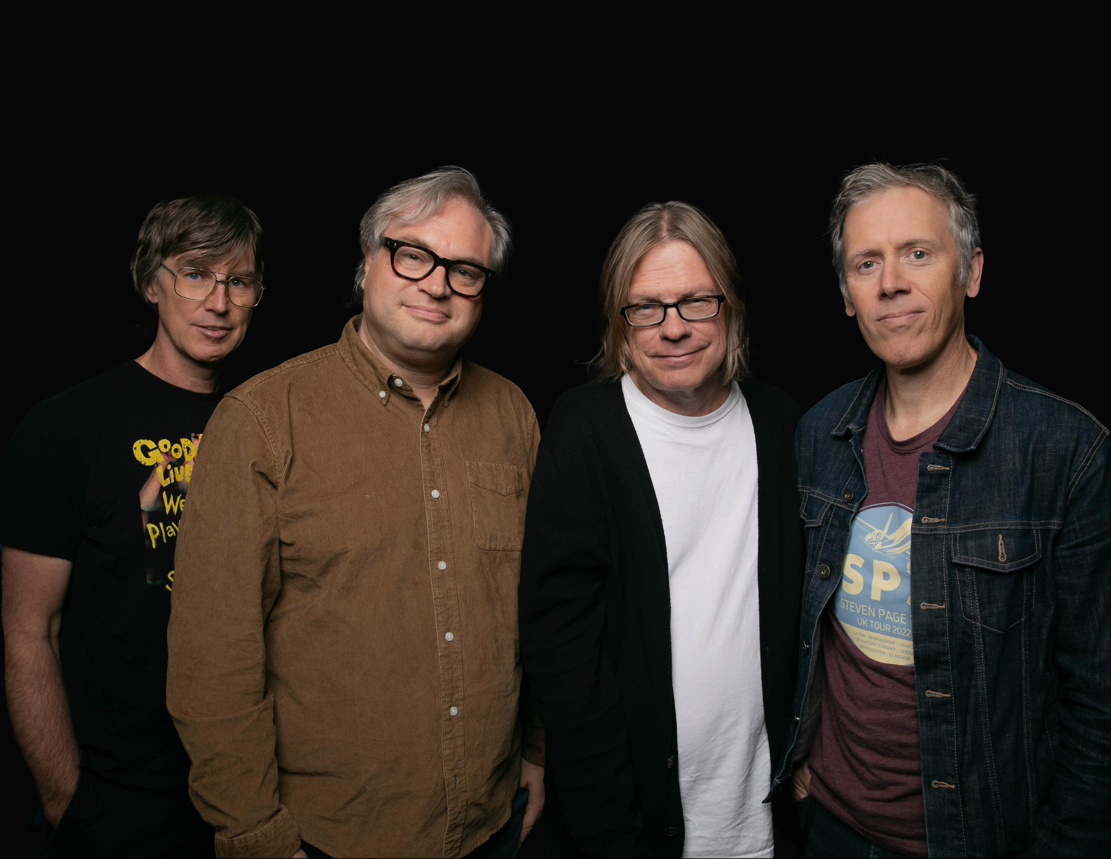 Four men stand in front of a black background and face the camera. The first man on the left is wearing a black t-shirt, the second man to the right wears a brown, long sleeved button-up shirt, the third man is wearing a white shirt covered by a black cardigan and the fourth man wears a maroon t-shirt with a black denim jacket on top.