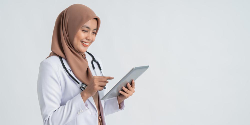 A smiling healthcare professional in a hijab uses a digital tablet.