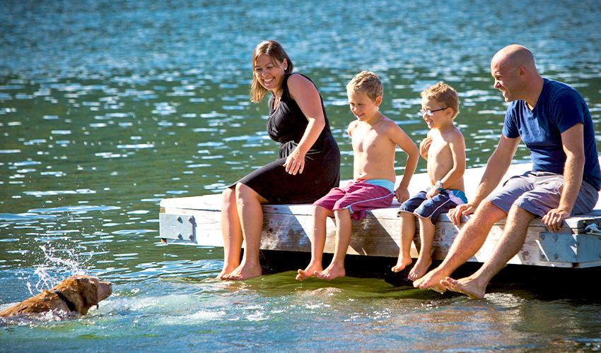 A family of four sitting on a dock on the water, there is a dog in the water swimming.