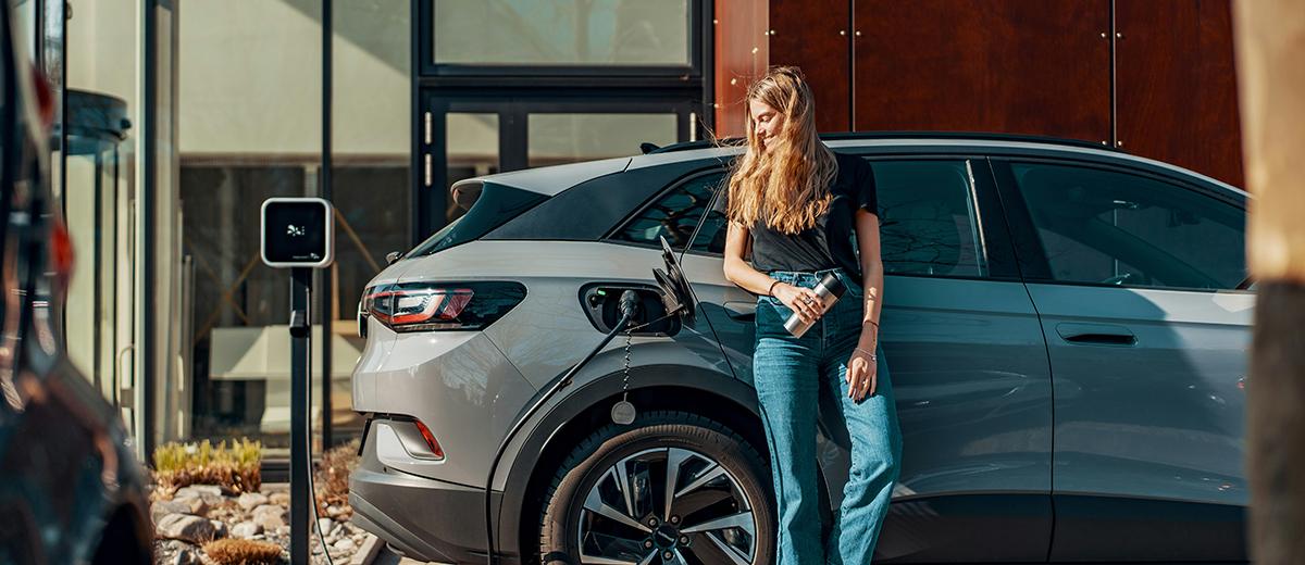 A woman stands to the side of her electric vehicle, leaning on the door as she watches it charge via the plugged in charger.