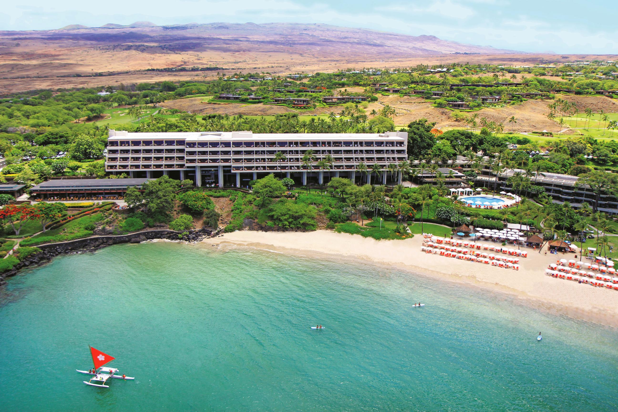 An overhead shot of a white, rectangular resort on a beach, surrounded by foliage. The water is bright blue and there is a small boat with a red flag just off the shore.