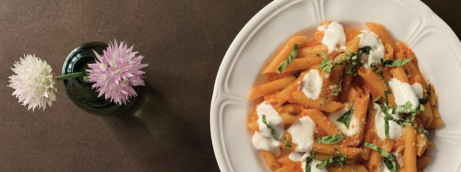An overhead shot of penne pasta in an orange sauce with bits of white cheese scattered throughout the dish. The place sits on a brown surface and, to the left, there is a green vase with two lilac flowers in it.