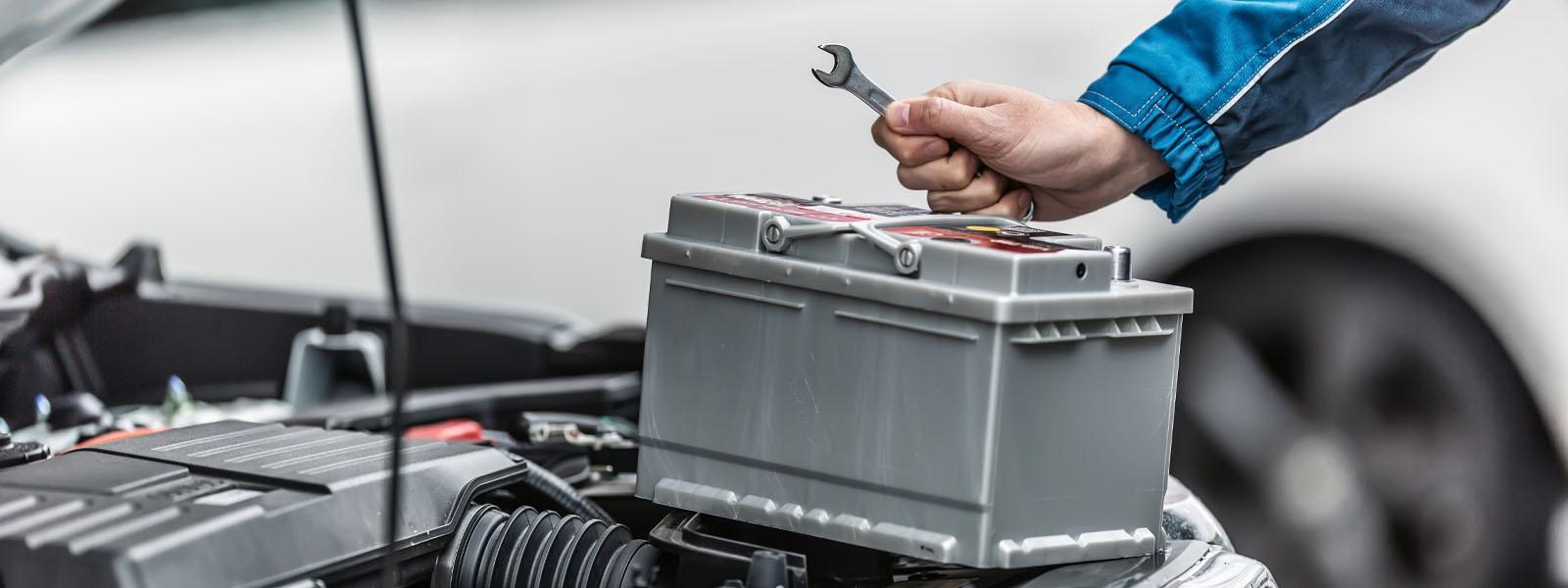 A vehicle’s hood is propped open and a battery is sitting on top of the engine with a mechanic’s hand holding a wrench above it. A white car is blurred in the background.