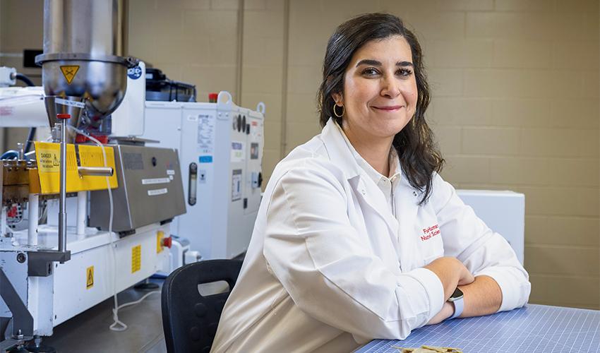 Food scientist smiling in a laboratory with food processing equipment in the background.