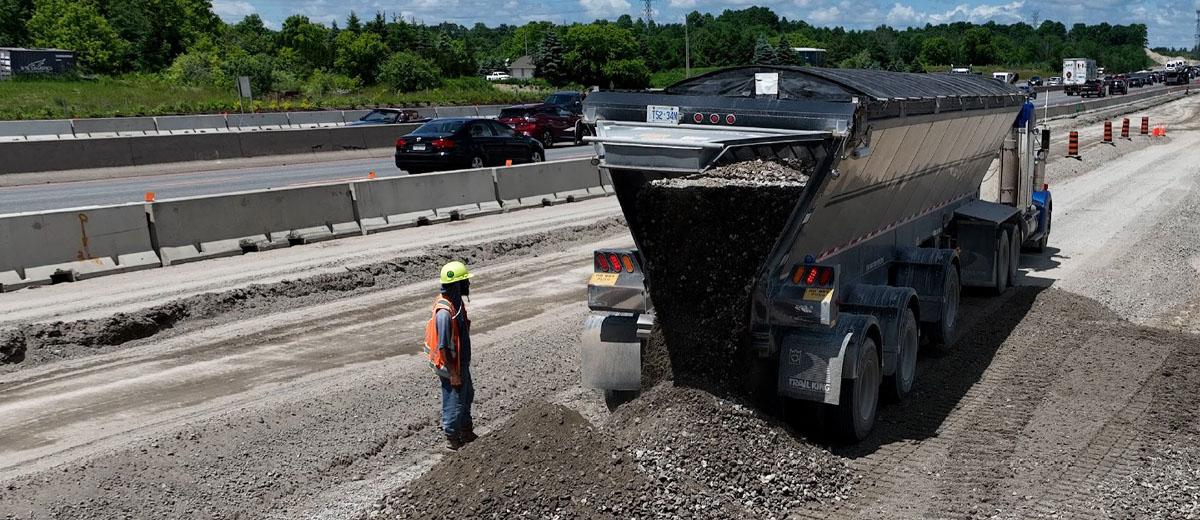 A construction worker stands on the side of a road, watching as a truck unloads asphalt onto the ground.