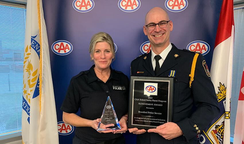Two individuals in uniform holding awards and smiling for the camera against a backdrop with banners including the caa logo.