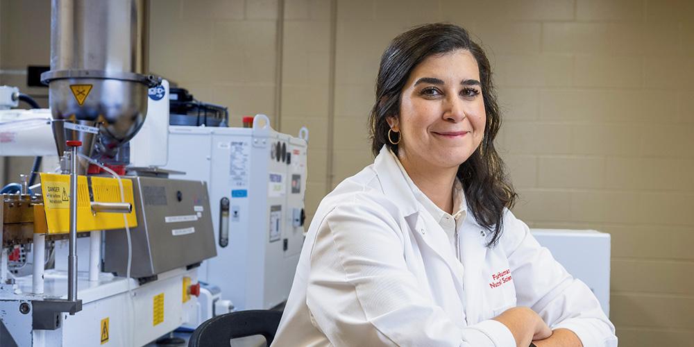 A woman scientist in a lab coat smiling at the camera with laboratory equipment in the background.