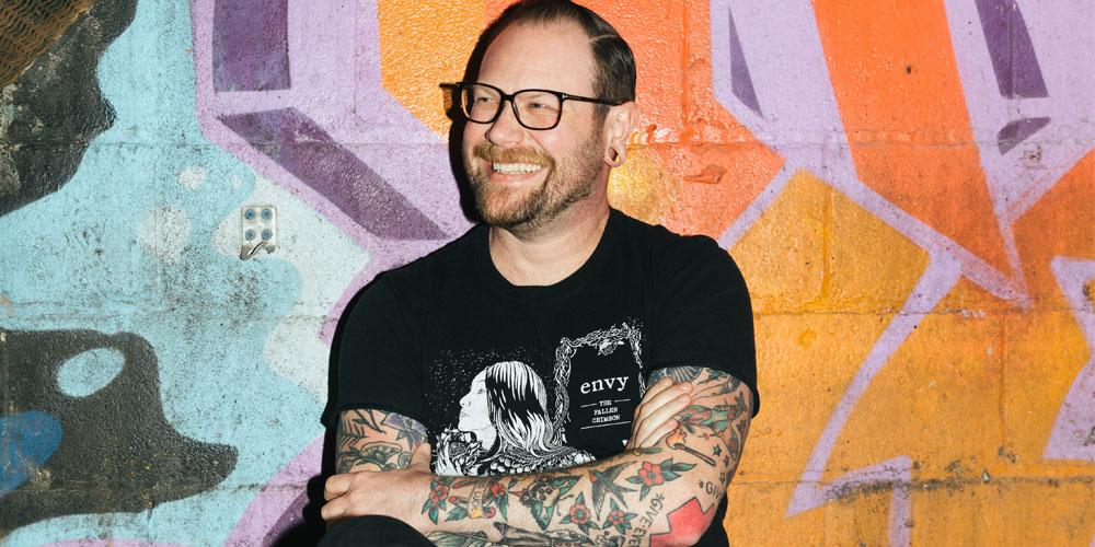 A man with tattoos sitting in front of a colorful wall.