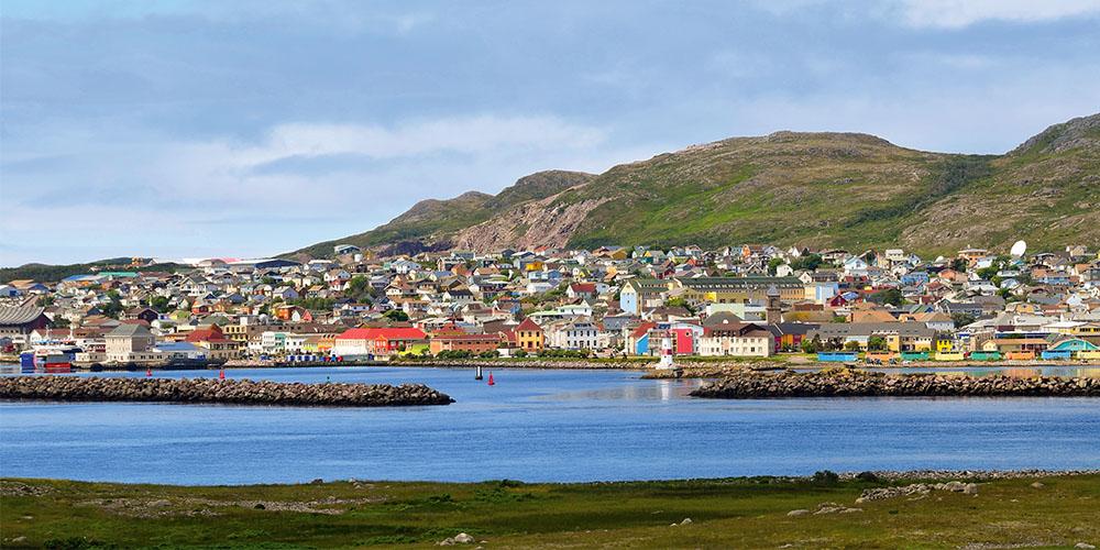 A colorful coastal town with houses and buildings against a backdrop of hills and a clear sky.
