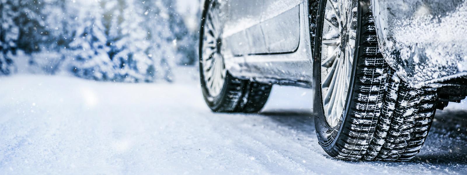 The lower half of the side of a vehicle, showing two of the vehicle's tires covered in snow. The road is also covered in snow, and in the distance are evergreen trees.