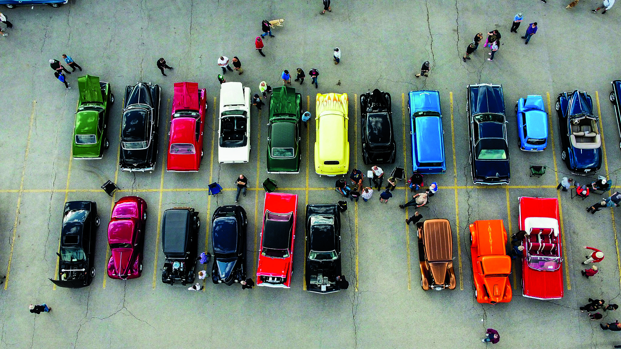 An overhead shot of classic cars of various colours, makes and models in a parking lot, with people surrounding them.