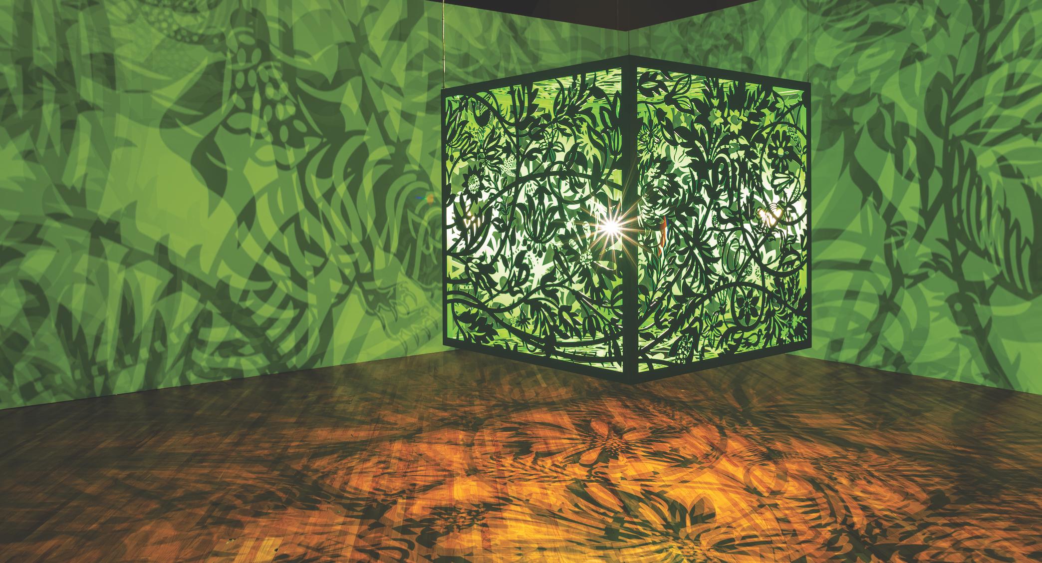 A box with black borders and sides etched with ornate designs floats in the middle of a room with green walls and a brown floor. The light omitted from the box and through the designs projects leaf-like shadows on the room.