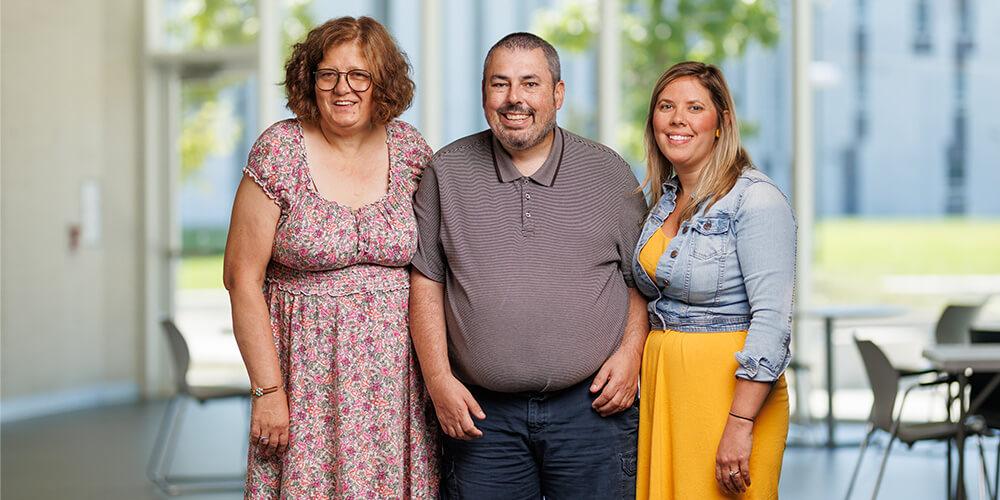 Three smiling adults standing together inside a building with windows and tables in the background.