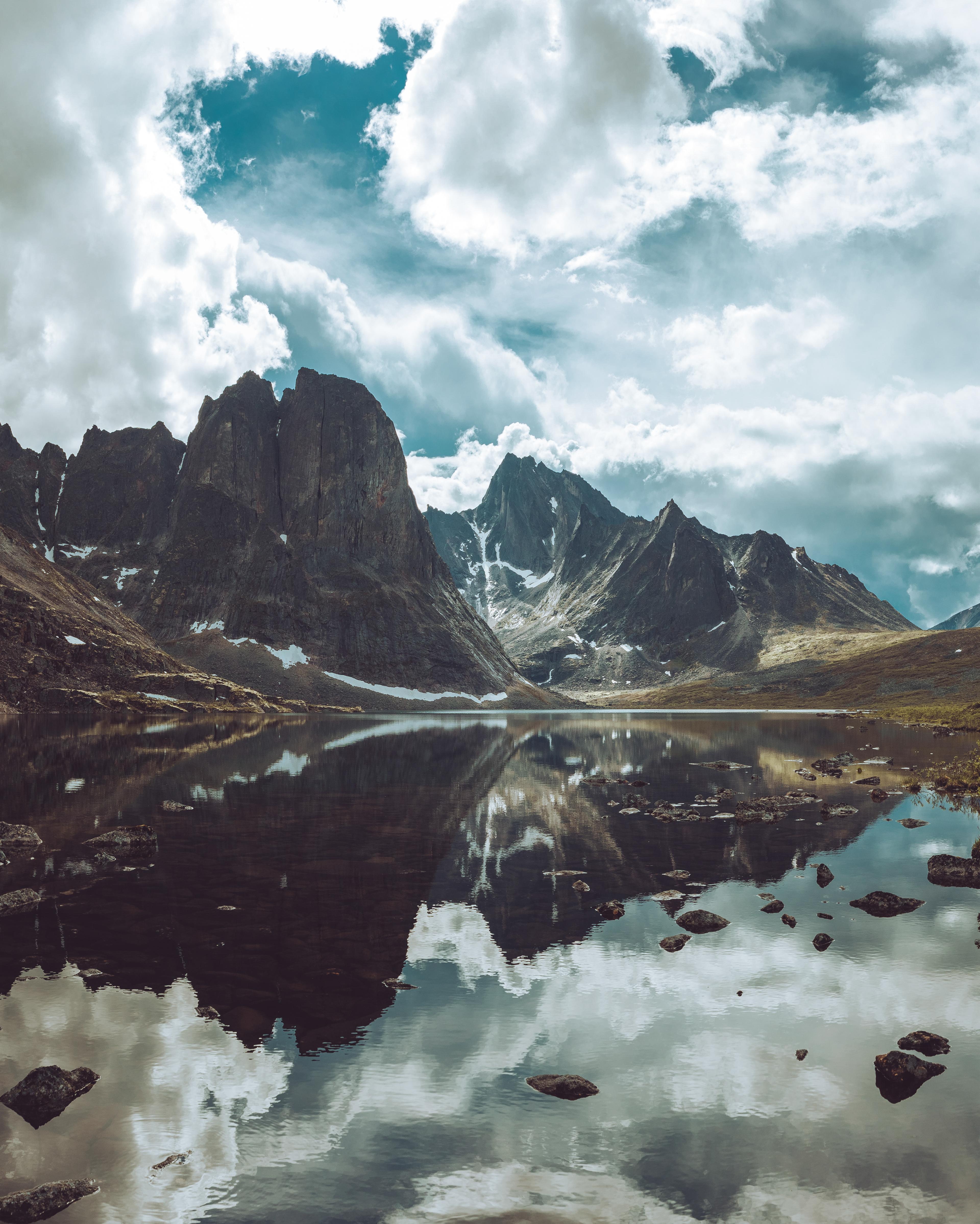 An image of a lake reflecting the large, dark grey mountains behind it, beneath a cloudy blue sky.