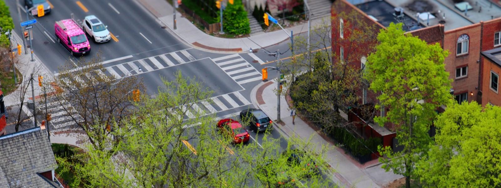 Aerial view of a vibrant urban intersection during daytime, featuring clearly marked pedestrian crosswalks, traffic signals, and several vehicles including a distinctive pink van with white graphics. The streets are lined with green trees and bordered by residential and commercial buildings. Sidewalks are well-maintained, and the scene reflects a blend of city infrastructure and natural elements, suggesting spring or early summer.