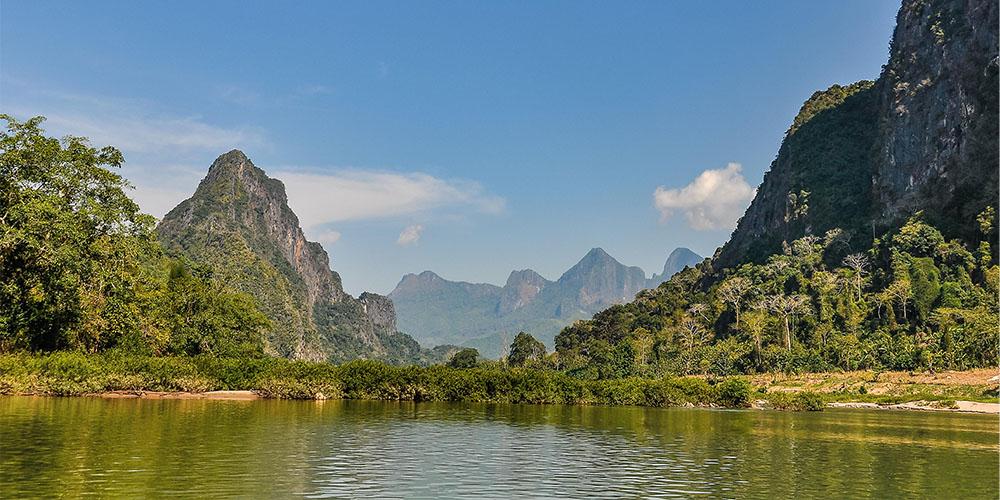 Lush riverside with towering karst mountains under a clear blue sky.