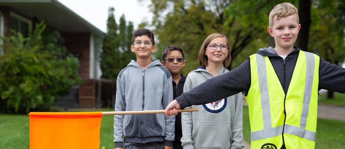 A child wearing a neon yellow vest and holding an orange flag stands with his arms outstretched, stopping the group behind him from crossing the street until it's safe to do so.