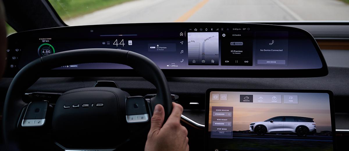 The interior of an electric vehicle; a hand holds the steering wheel. Behind the wheel are various screens showing different information about the car and the route.