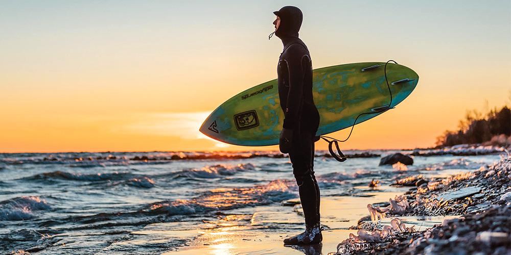 A man in a wetsuit holding a surfboard at sunset.