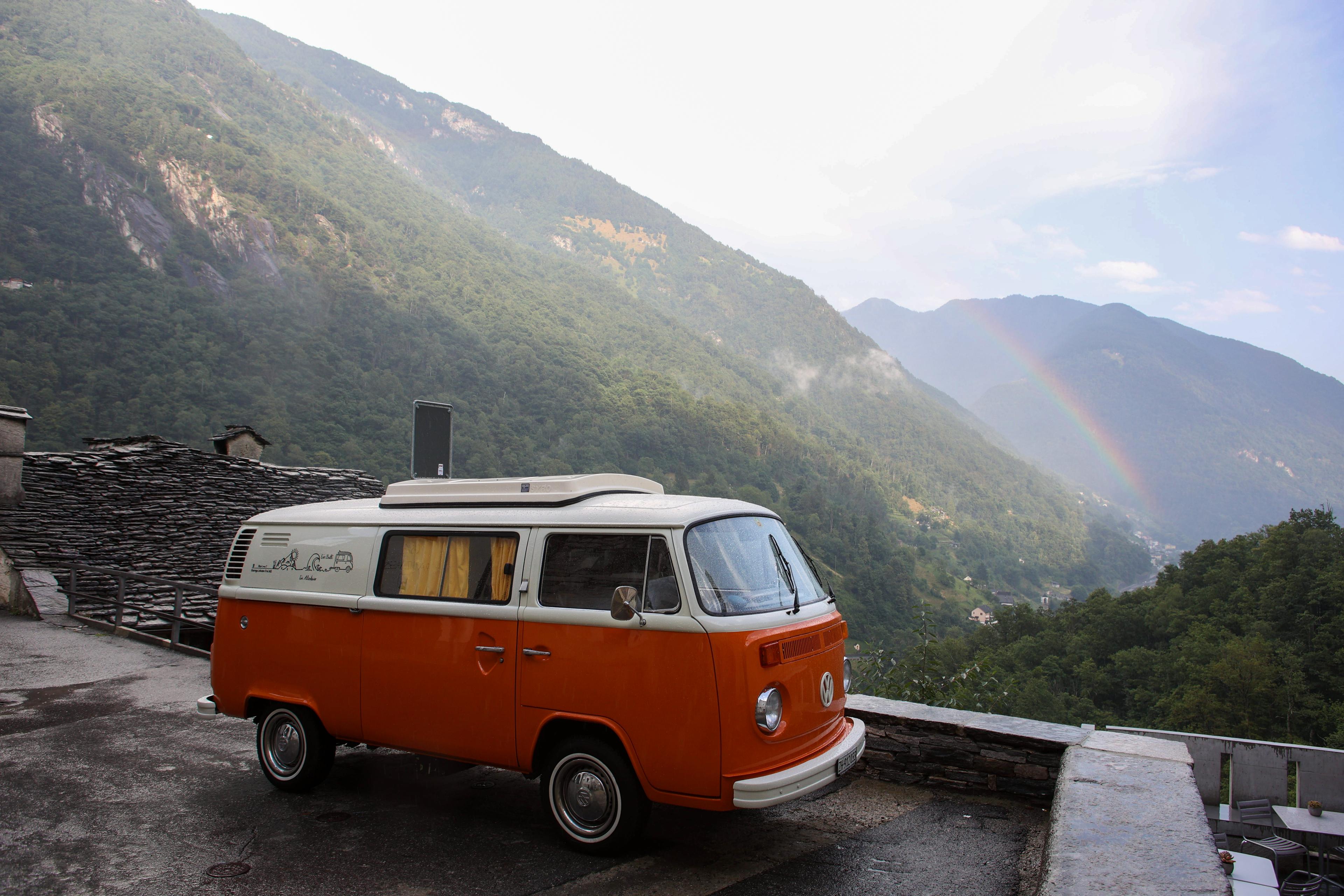 A vintage red and white Volkswagen parked on a road with views of mountains behind it. The sky is clear and there is a small rainbow in the distance.
