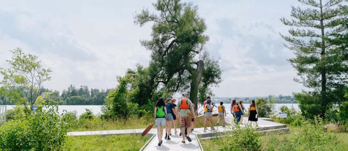 On a warm sunny day, a group of people in life jackets are walking along a wooden path down the water, some carrying paddles.
