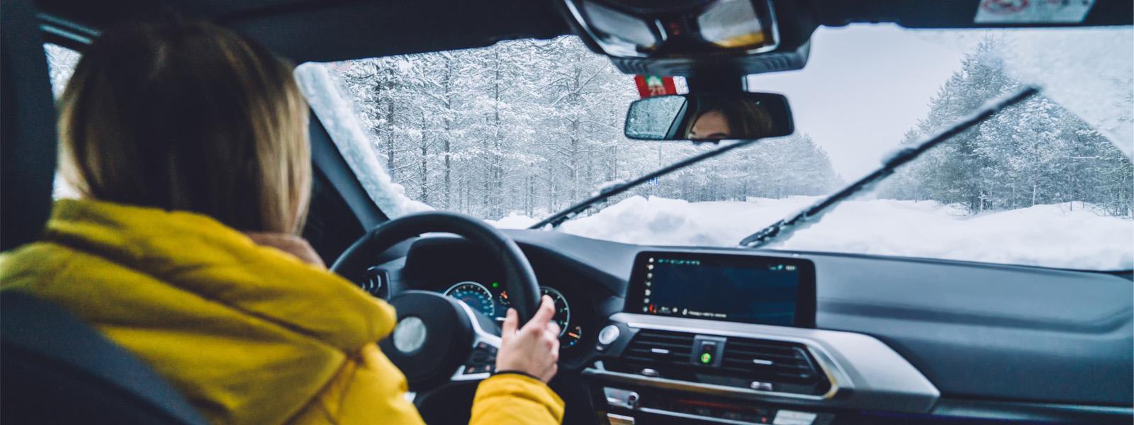 A woman wearing a yellow jacket with blonde hair sits in the driver's seat with her hands on the wheel. Through the windshield, it shows that she is driving through a forest in the winter, with snow covering the road in front of her.