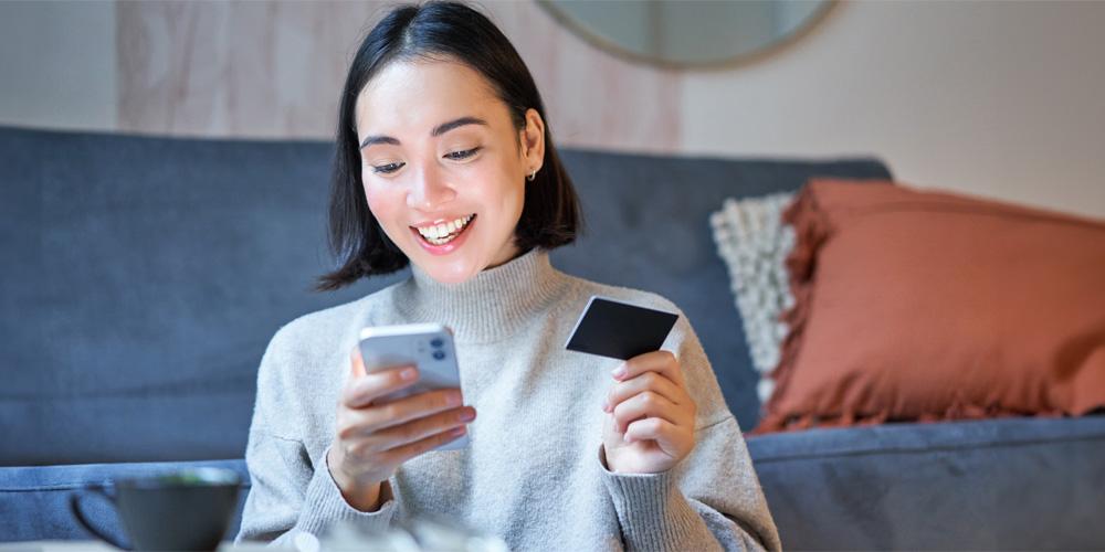 A woman smiling while looking at her smartphone and holding a credit card.