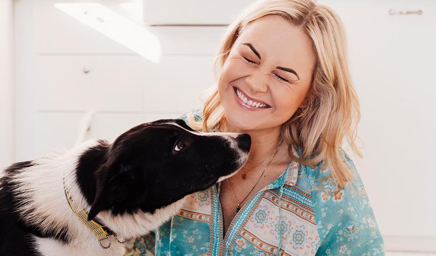 A woman smiles while petting her dog in the kitchen.