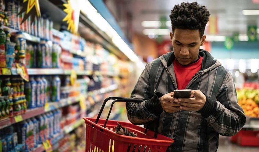 Male looking down at mobile phone and typing while in a grocery item, he has a grocery basket hanging from his arm.