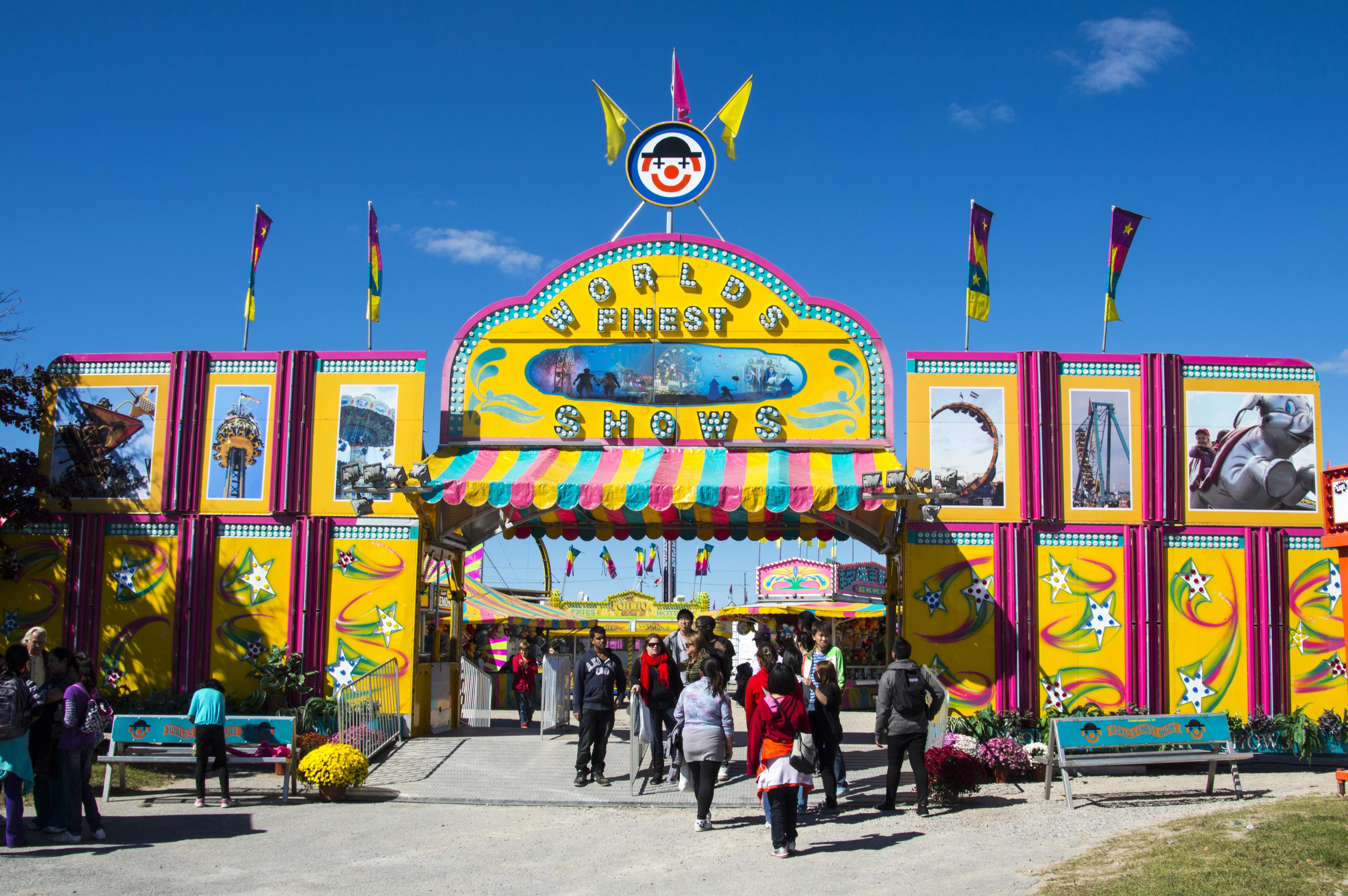 The entrance of a fair: a large, brightly coloured doorway that says "World's Finest Shows" above the walkway. The sky above it is blue and there are images of various fair rides plastered on the front.