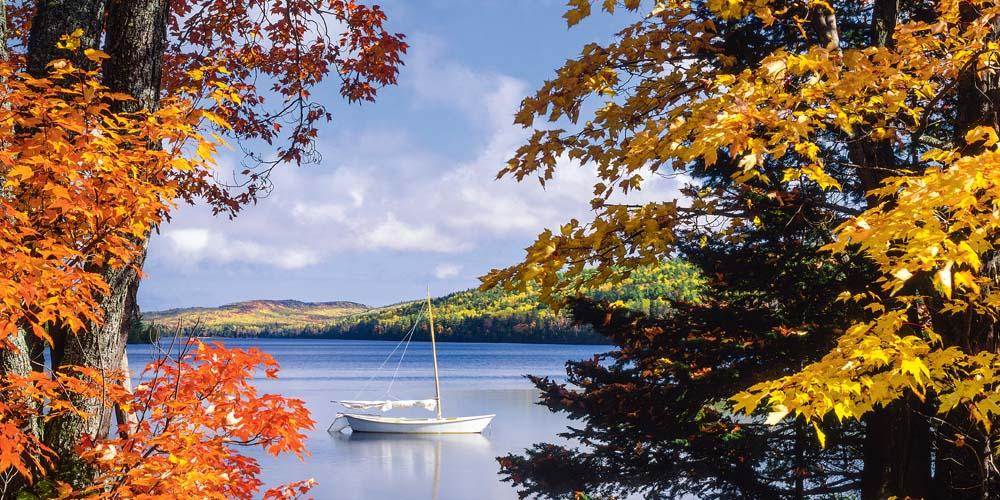 A sailboat on a calm lake framed by autumn-colored foliage.