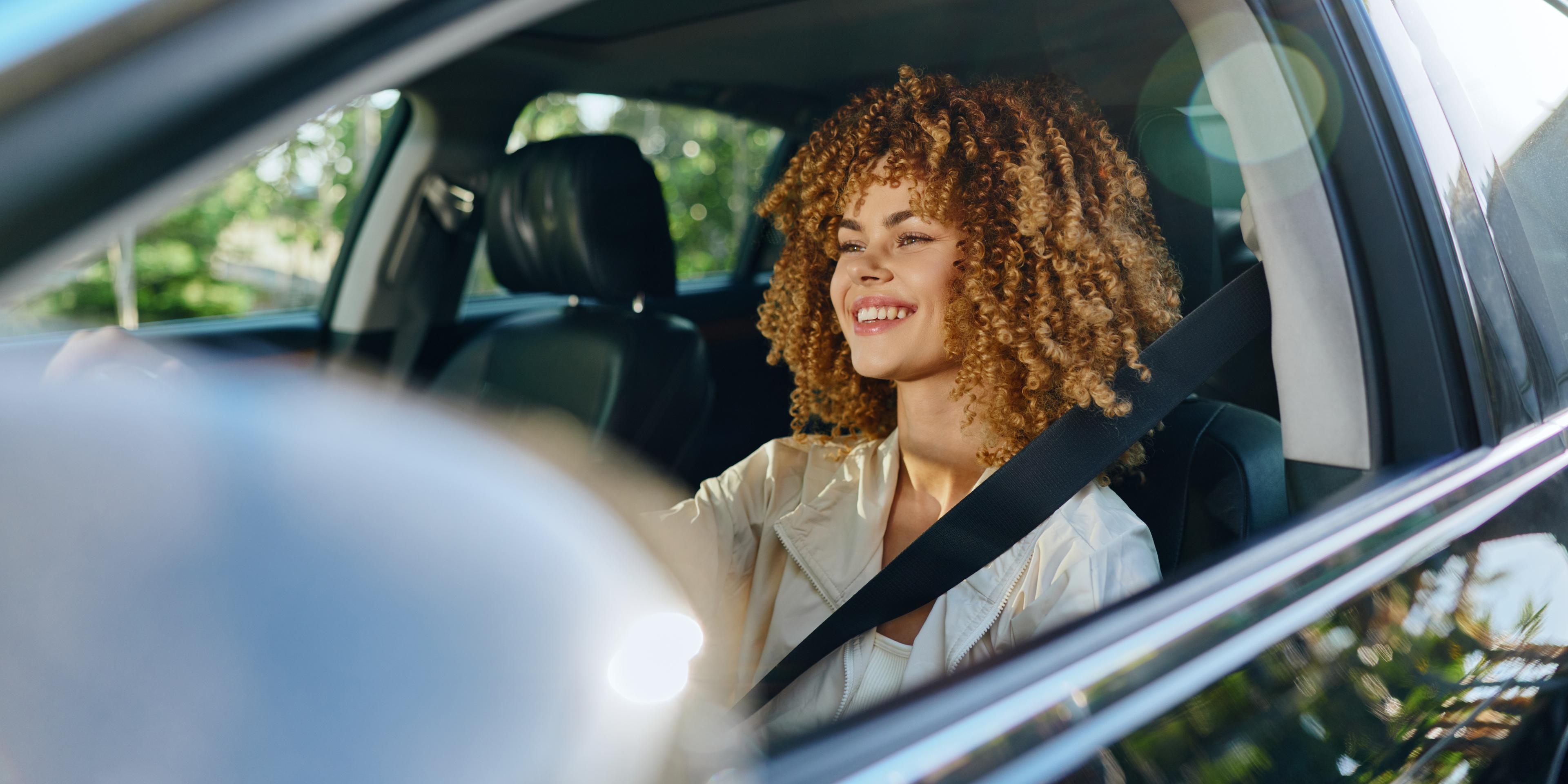 A woman sits in the driver's seat of a car with her seatbelt on; she is looking out at the road and smiling.