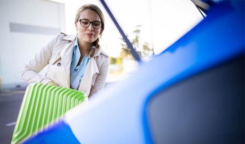 A woman is holding a blue suitcase in front of a blue car.