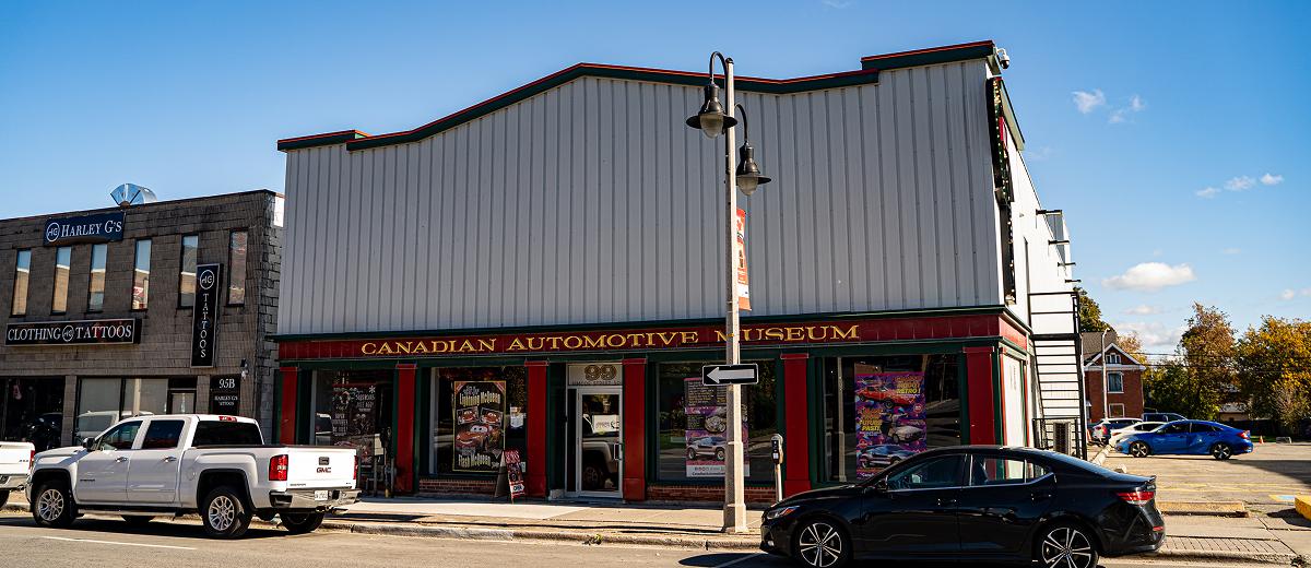 The exterior of the Canadian Automotive Museum, with a sign out front depicting the name. The windows are framed in red and green, while the top half of the building is made of grey panels.
