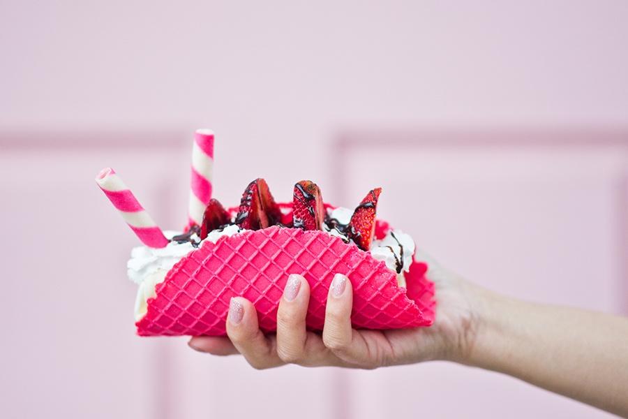 A hand holding a pink waffle cone with strawberries and whipped cream.
