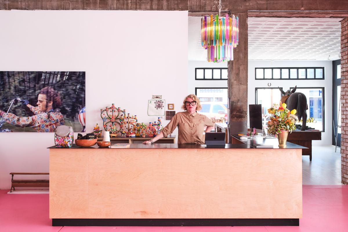 A woman stands behind a salmon-coloured desk in the lobby of an art gallery with pink carpet. Above her is a rainbow-coloured chandelier and behind the desk are various art pieces including a painting of a man on a motorcycle.