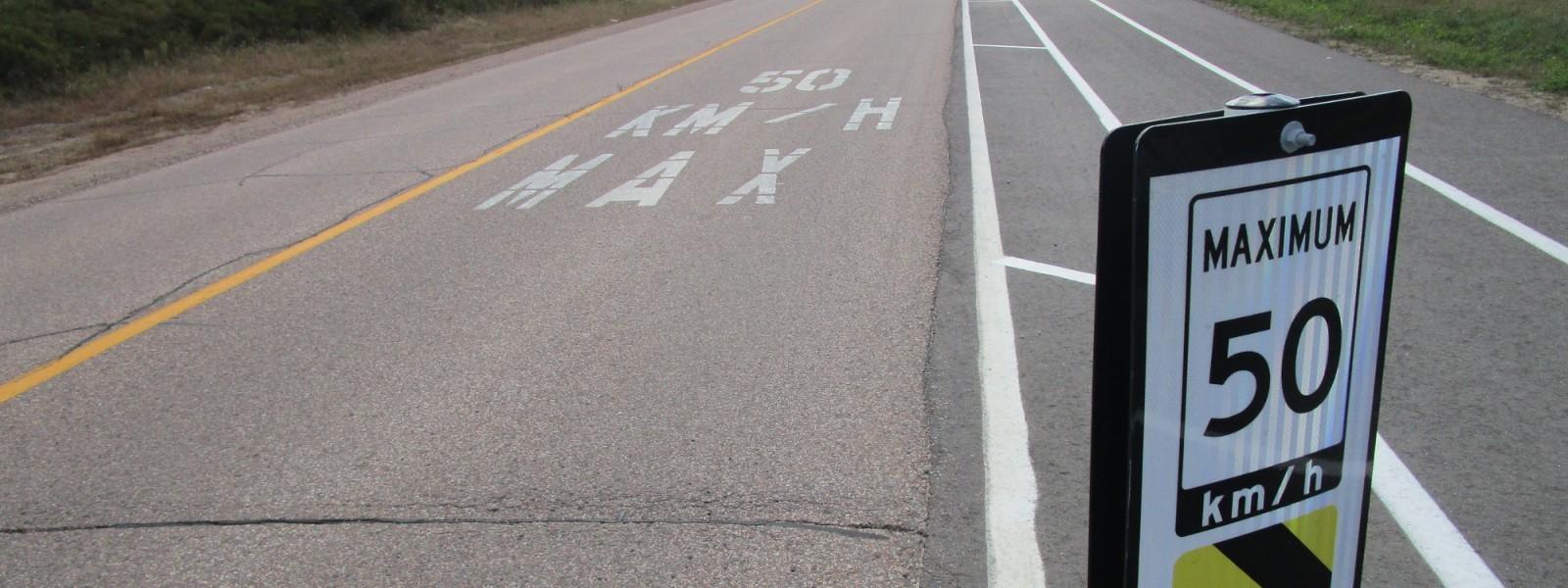 A multi-lane road with a posted speed limit sign reading 'MAXIMUM 50 km/h' and matching road markings that say '50 KM/H MAX' in large white letters. The road includes a bike lane or shoulder lane marked by white lines, emphasizing traffic regulations and safety measures.