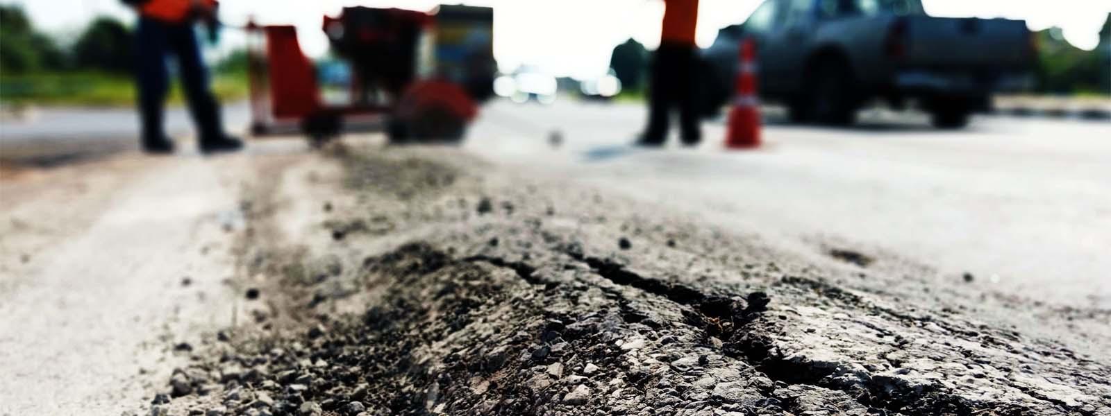 In the foreground there is a raised crack in the road. In the background, blurry is a parked pick-up truck and the outlines of construction workers working to repair the road.