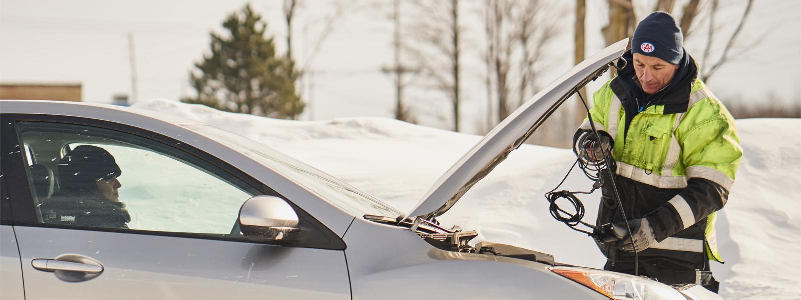 A CAA service vehicle operator, wearing a neon green jacket and a black CAA-branded toque, props up the hood of a car outside in the snow. Sitting in the driver's sear of the car is a woman wearing a black scarf and toque.